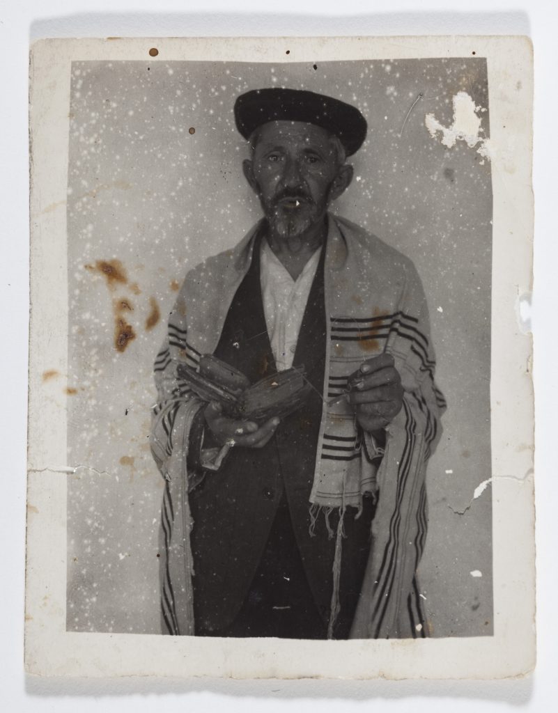 Das Foto zeigt einen älteren Mann mit nachdenklichem Gesichtsausdruck. Er trägt einen Gebetsschal und hält ein aufgeschlagenes Buch. Das Foto selbst hat starke Alterungsspuren.