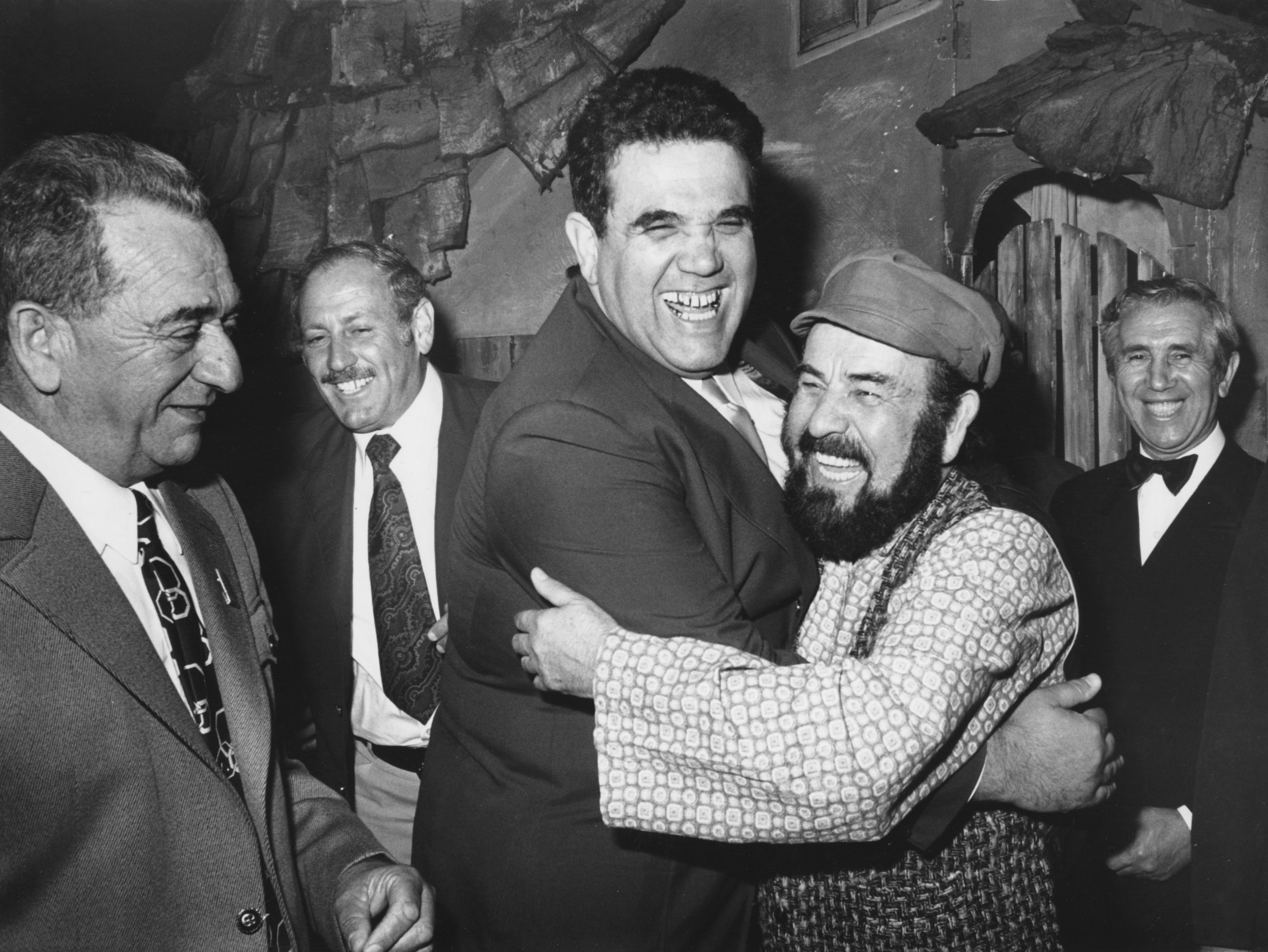 Yossef Gutfreund and Shmuel Rodensky hug each other and laugh. Around them stand three men, smiling broadly. All of them are wearing suits, Rodensky a patterned wide shirt and a cap. Backdrops can be seen in the background.
