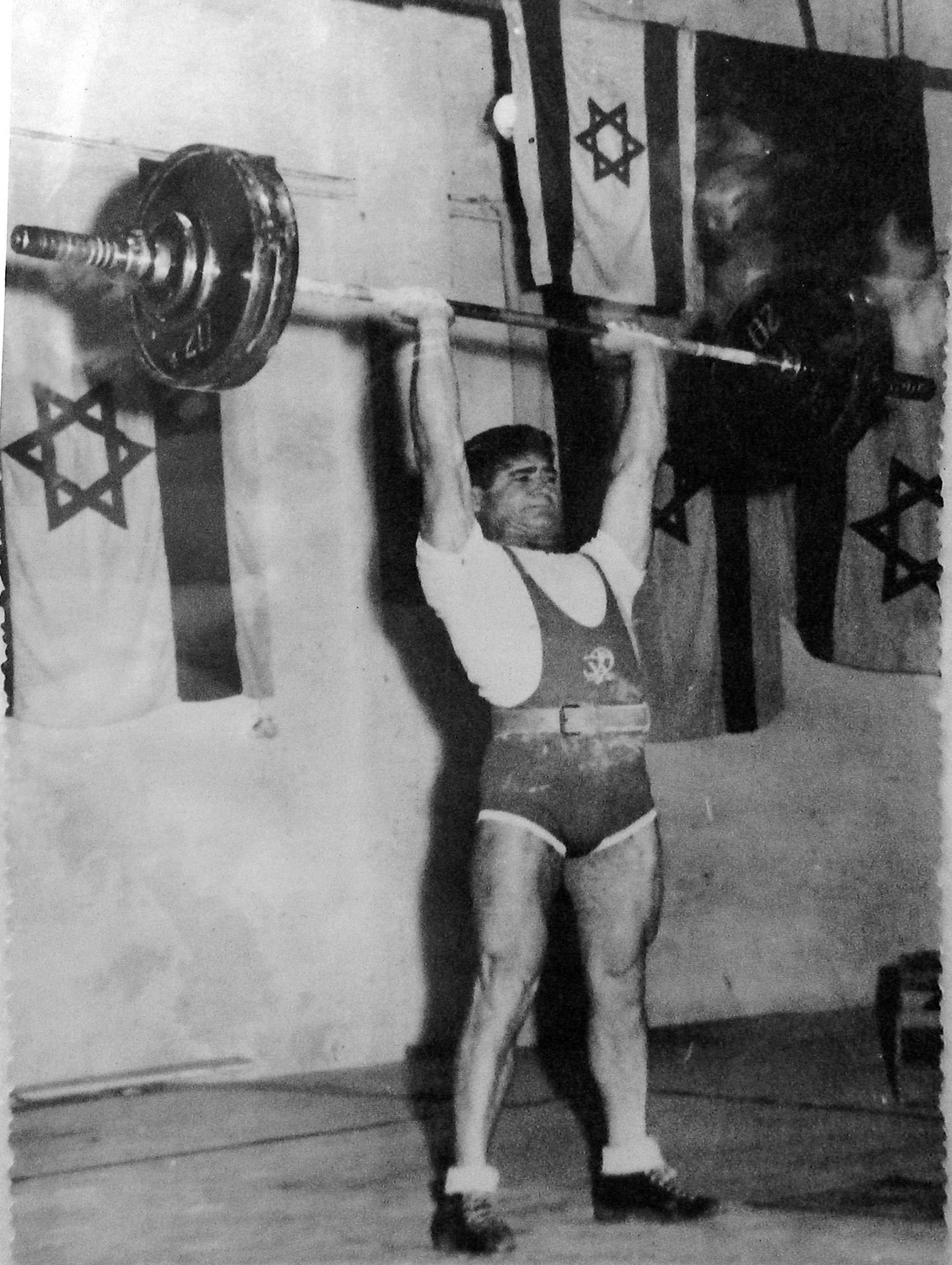 Romano stemmt ein Gewicht in die Höhe. Im Hintergrund sind Israelflaggen.
