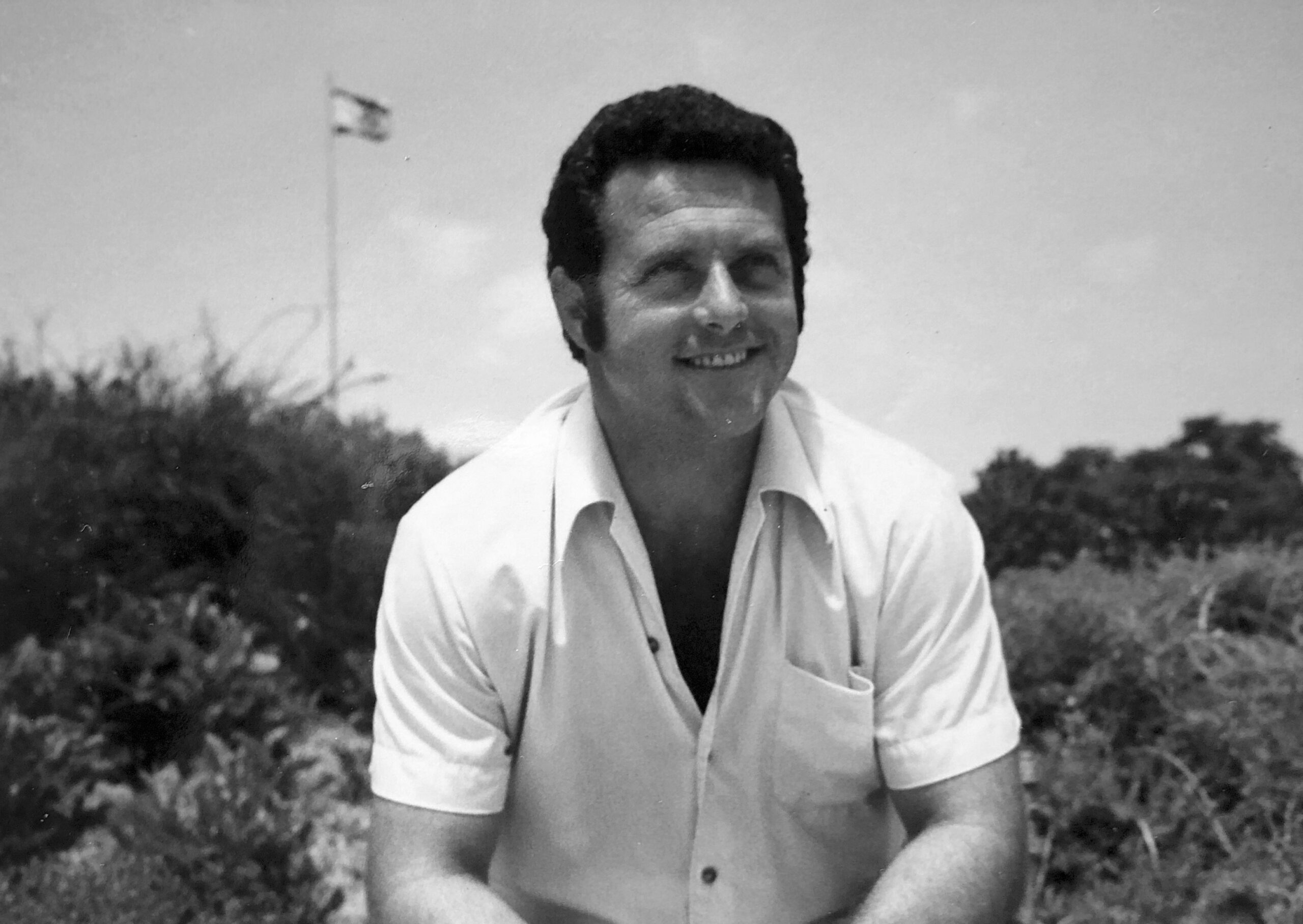 Portrait Moshe Weinberg im Freien. Er trägt ein helles kurzärmeliges Hemd und lacht. Im Hintergrund sind Sträucher und ein Fahnenmast mit israelischer Flagge zu sehen.