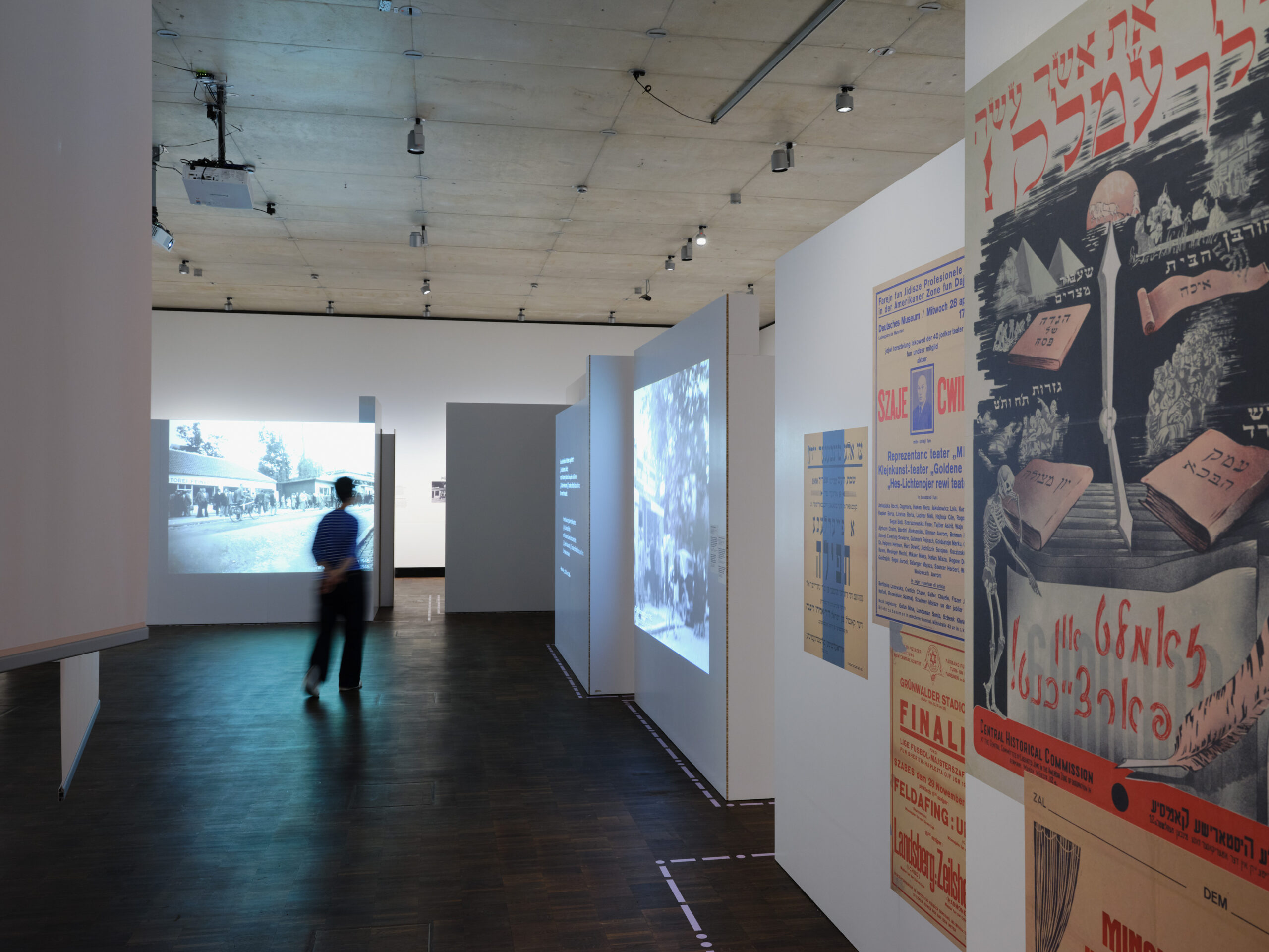 Ausstellungsansicht mit Besucherin, rechts ist eine Wand mit verschiedensprachigen Plakaten zu erkennen - einige in Jiddisch; links hinten ein Schwarz-Weiß-Video