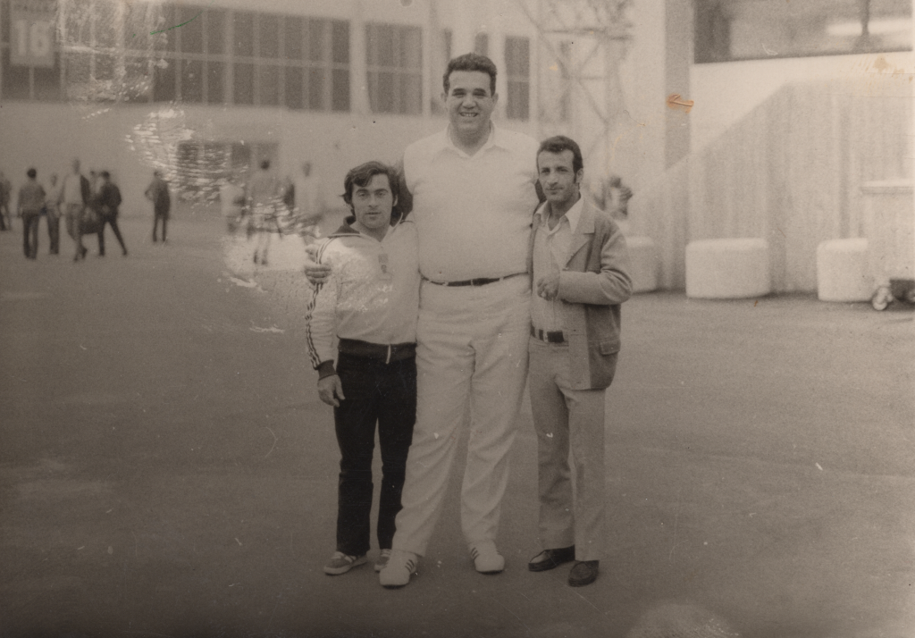 Three men in athletic clothing stand in front of a hall. Yossef Gutfreund in the middle puts his arms around the other two. He is two heads taller than the shorter of the two.  
