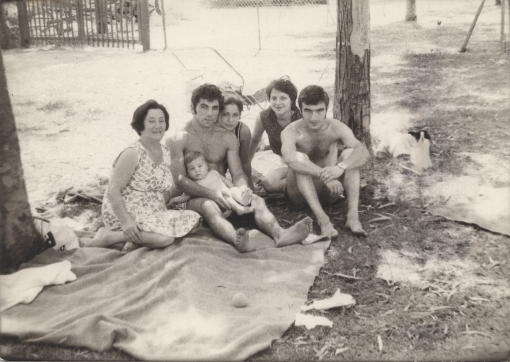 Gruppenfoto, schwarz-weiß: Die Familie sitzt im freien auf einer Picknickdecke. Die Männer sind nur in Schorts, die Frauen in sommerlichen Kleidern. 