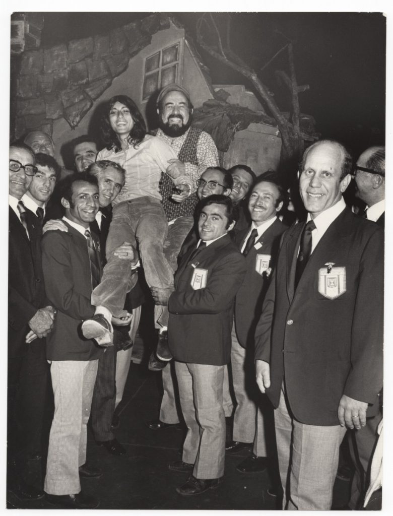 Members of the Israeli Olympic team stand on the stage and lift Esther Shahamorov and Shmuel Rodensky into the air. Everyone laughs exuberantly into the camera. 