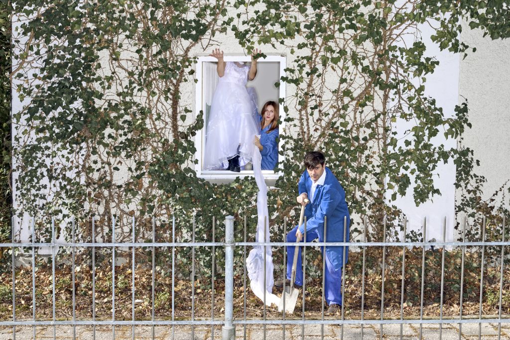 Christoph Theussl steht mit einem Spaten vor einem mit Efeu bewachsenen Haus. Judith Huber und eine Person in einem Brautkleid, deren Gesicht nicht zu erkennen ist, stehen im geöffneten Fenster. Judith und Christoph tragen blaue Arbeitsanzüge.