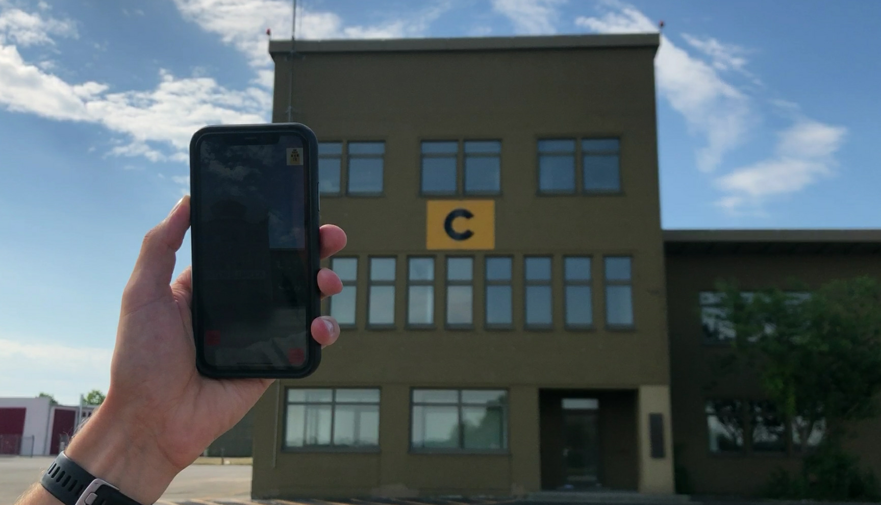 Eine Person hält ein Handy in die Luft, im Hintergrund ist ein Gebäude des Fliegerhorsts Fürstenfeldbruck zu erkennen.