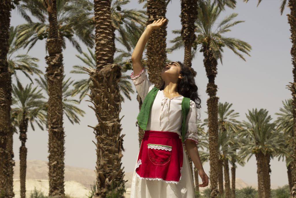 Junge Frau in Trachtenkostüm steht unter Palmen und hält ihren Arm vors Gesicht, um sich vor der grellen Sonne zu schützen.