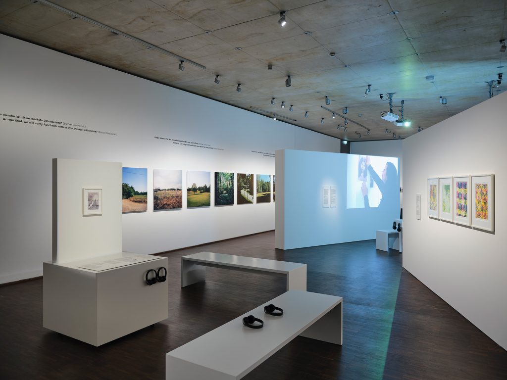 Ausstellungsansicht mit Fotografien und Drucken an der Wand sowie einer Videoprojektion und Sitzbänken mit Kopfhörern