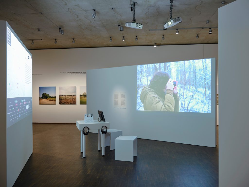 Installationsansicht mit einer Videoprojektion von einer jungen Frau mit Kamera und einem Tisch mit Kopfhörern
