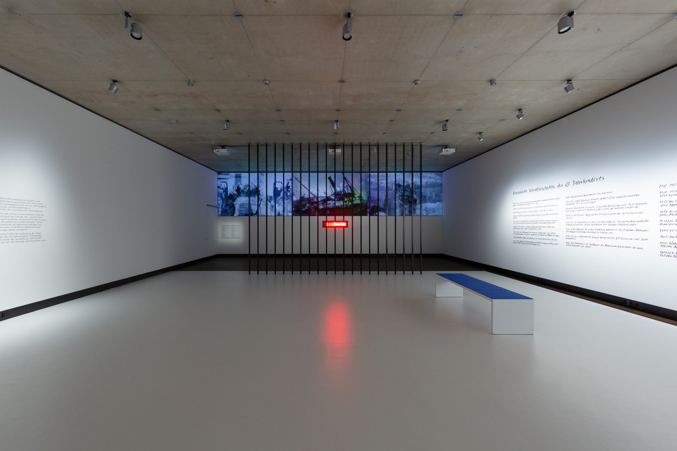 Blick in den Ausstellungsraum: Vor einer Videoprojektion sind deckenhohe schwarze Gitterstäbe, an denen eine rote Leuchtanzeige mit einer Zahl montiert ist. An den Wänden rechts und links Text, davor eine Bank.
