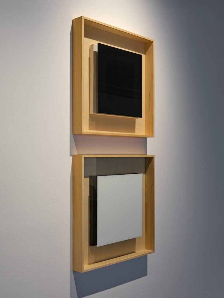 Auf dem Foto sind zwei quadratische Holzbilderrahmen übereinander zu sehen. In dem oberen Rahmen hängt ein quadratisches Bild, das komplett schwarz ist. Das untere Bild ist komplett weiß.