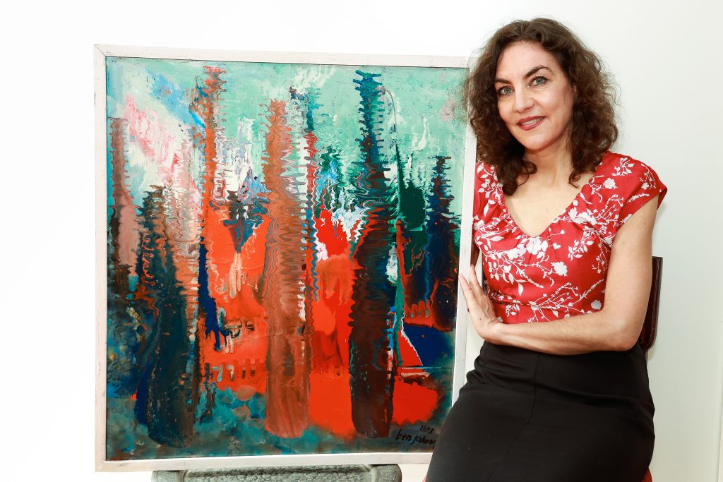 Judith Faessler hält ein Gemälde mit roten, blauen, schwarzen und braunen verschwommenen Lilien und Mustern in ihrem Arm.