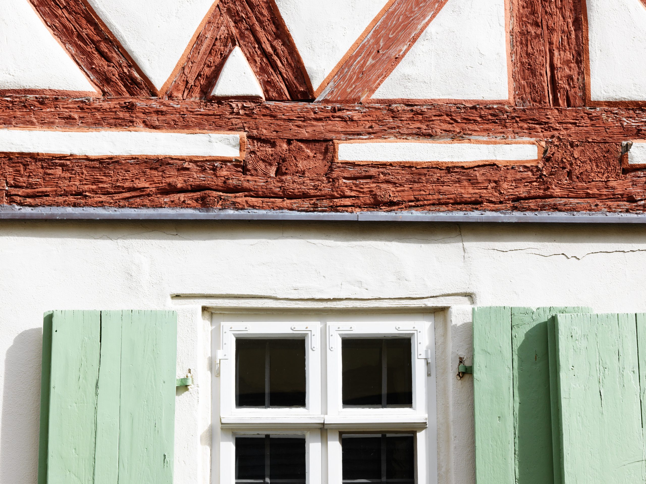 Detailaufnahme, Fachwerkfassade mit Fenster. Oben braune Holzbalken und weißer Putz, unten ein Fenster mit geöffneten hellgrünen Fensterläden.