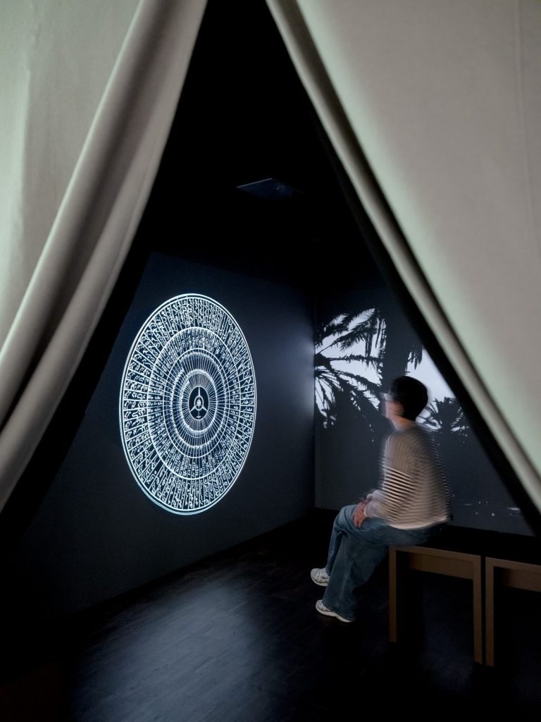 Hinter einem geöffneten Vorhang sitzt eine Frau auf einer Bank und schaut auf eine Beamer Installation an der Wand. Es ist ein großes Mandala. Rechts auf dem Bildschirm hinter der Frau sind Palmen von einer anderen Video Installation zu sehen.