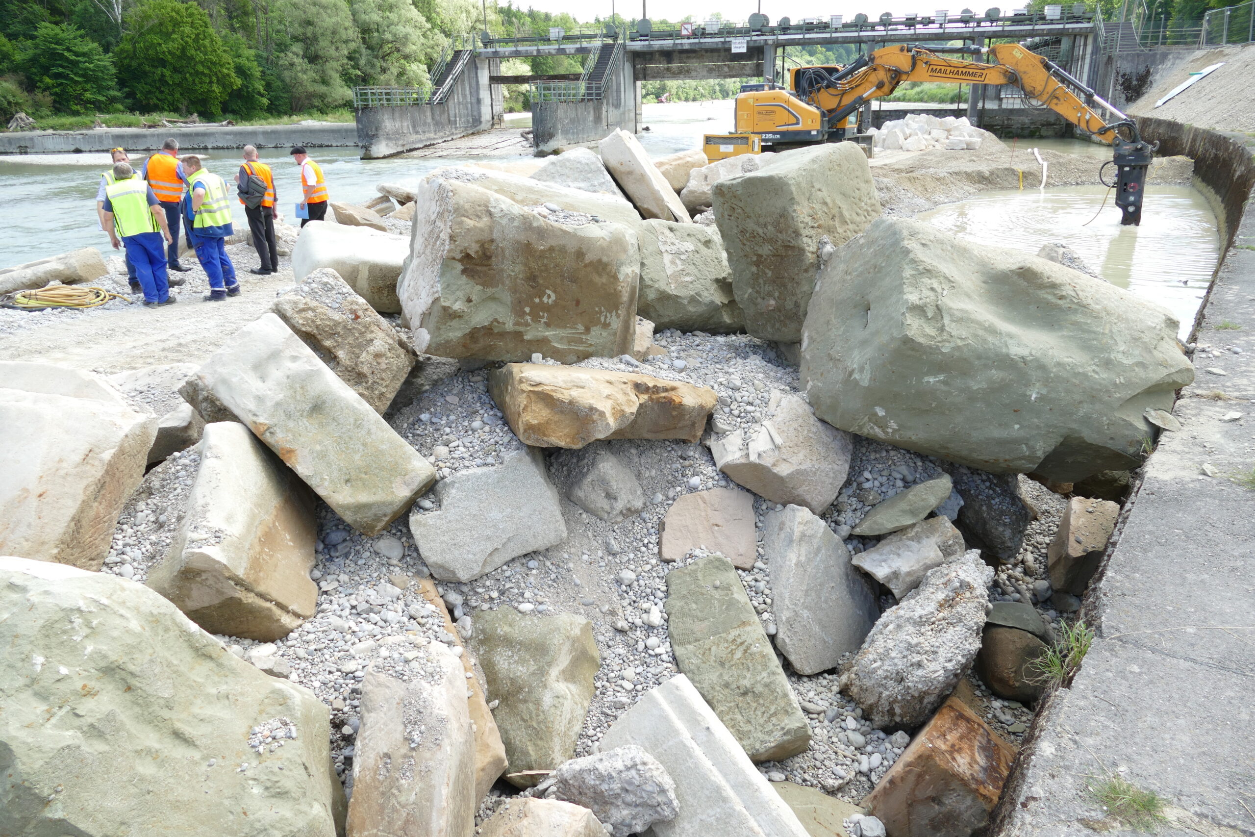 Flussufer mit aufgetürmten Steinen. Im Hintergrund sind ein Bagger und mehrere Menschen in Warnwesten