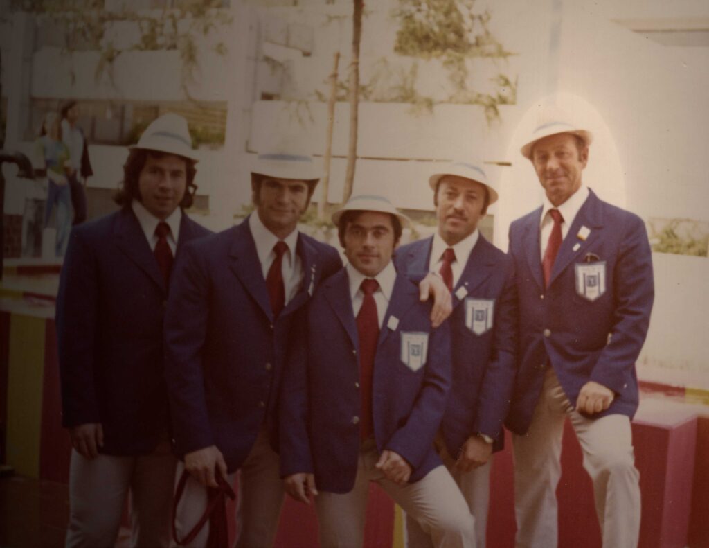 Group photo: Five men in the suits of the Israeli delegation with badges and hats. The Olympic Village is visible in the background.