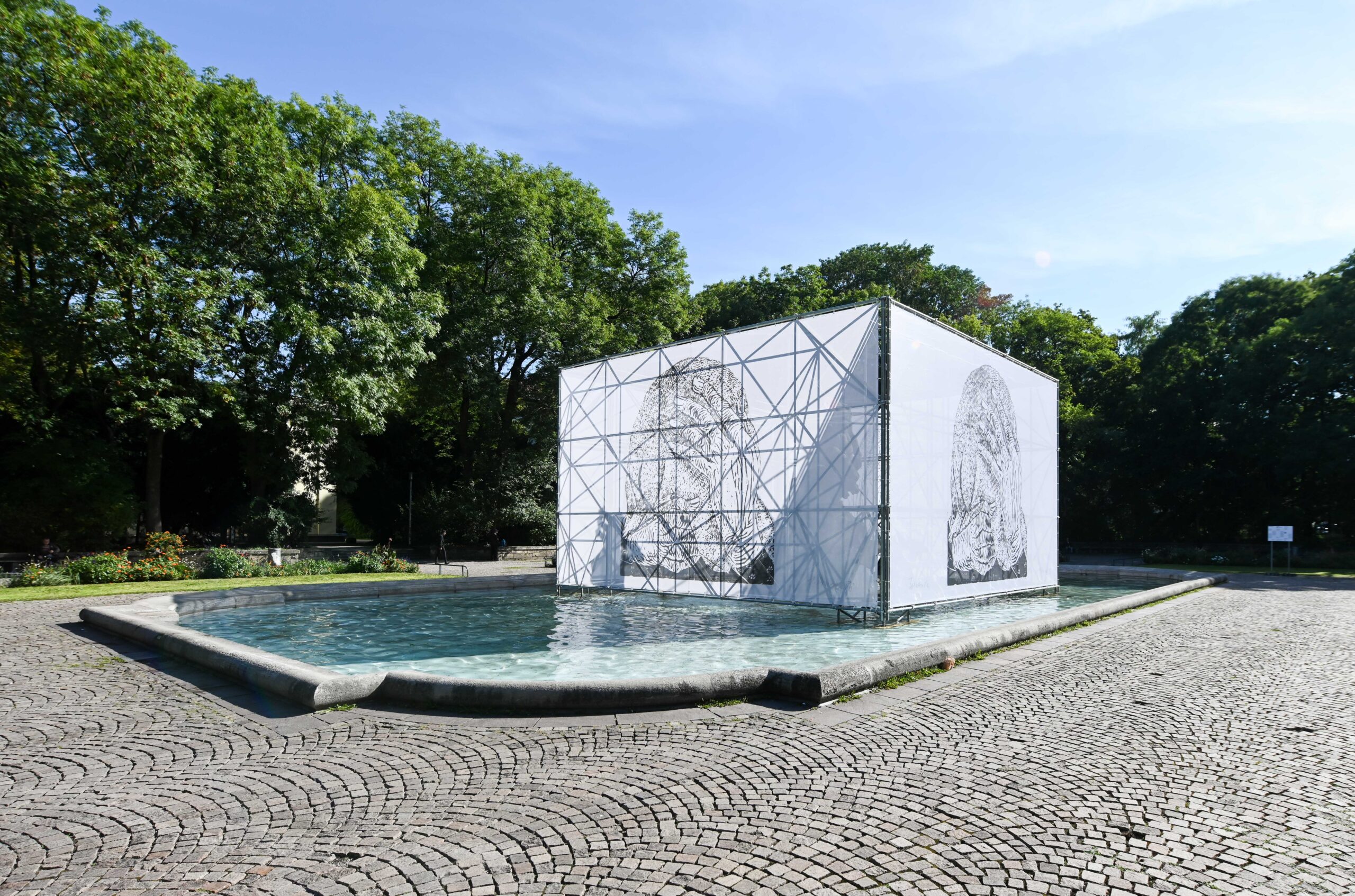 Platz mit Brunnen und Bäumen. Die Brunnenfigur ist von einem Gerüst mit einer bedruckten Folie verdeckt. Auf der Folie ist der Holzdruck „Trauernde“ von Maria Luiko zu erkennen, eine Figur in Schwarz-Weiß, die ihren Kopf mit einem Tuch verdeckt. Sonne scheint durch die Folie.