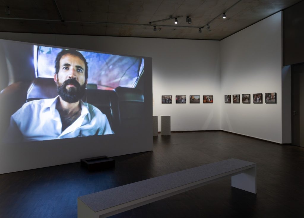 Auf dem Foto ist ein Ausschnitt aus der Ausstellung zu sehen. Links ist auf einer Wand ein großes Beamer Bild, das einen Film zeigt. In der Sequenz ist ein schwarzhaariger Mann mit dichtem Bart auf dem Rücksitz eines Autos zu sehen. Vor dem Bild steht eine Bank. Rechts von der Filmszene sind an einer Wand mittig mehrere Bilder in rechteckigen Bilderrahmen zu sehen.