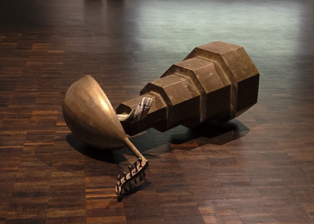 Auf dem Bild ist ein Metallobjekt, das auf dem Boden liegt, zu sehen. Der rechte Teil sieht aus wie eine Torte mit mehreren kleiner werdenden Ebenen. Obendrauf steckt ein Musikinstrument und ein Widderhorn.