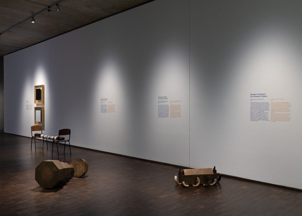 Auf dem Foto ist ein Ausschnitt der Ausstellung zu sehen. Vor einer großen weißen Wand mit Textfeldern liegen Metallobjekte auf dem Boden. Das rechte Objekt sieht aus wie eine Truhe. Daran sind mehrere Hörner befestigt. Links im Bild sind zwei weitere Kunstwerke des Künstlers zu sehen.