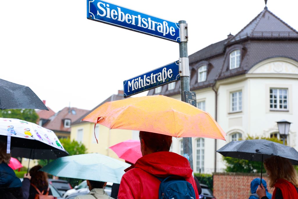 Menschen mit bunten Regenschirmen laufen an den Straßenschildern "Möhlstraße" und "Siebertstraße" vorbei