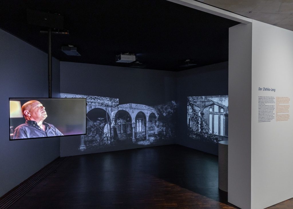 Auf dem Foto ist ein Raum der Ausstellung zu sehen. Links hängt ein Bildschirm, auf dem ein Herr mit Glatze in Hemd zu sehen ist. Rechts hinter im werden auf zwei Wände Videos einer Synagoge gezeigt.