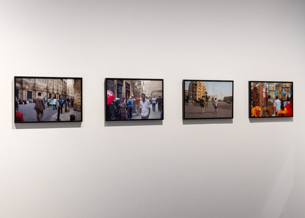 Auf dem Foto sind vier schwarze Bilderrahmen auf einer weißen Wand zu sehen. Auf den Fotos erkennt man Personen in einer städtischen Szenerie.