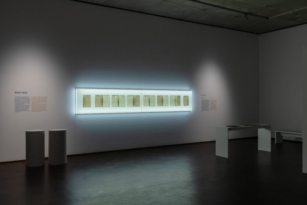 Auf dem Foto sieht man einen Ausschnitt der Ausstellung. An einer weißen Wand hängt mittig eine lange rechteckige Vitrine. Sie leuchtet, in ihr befinden sich mehrere DIN4 Seiten. Links und rechts vom Kunstwerk ist Wandtext zu sehen.