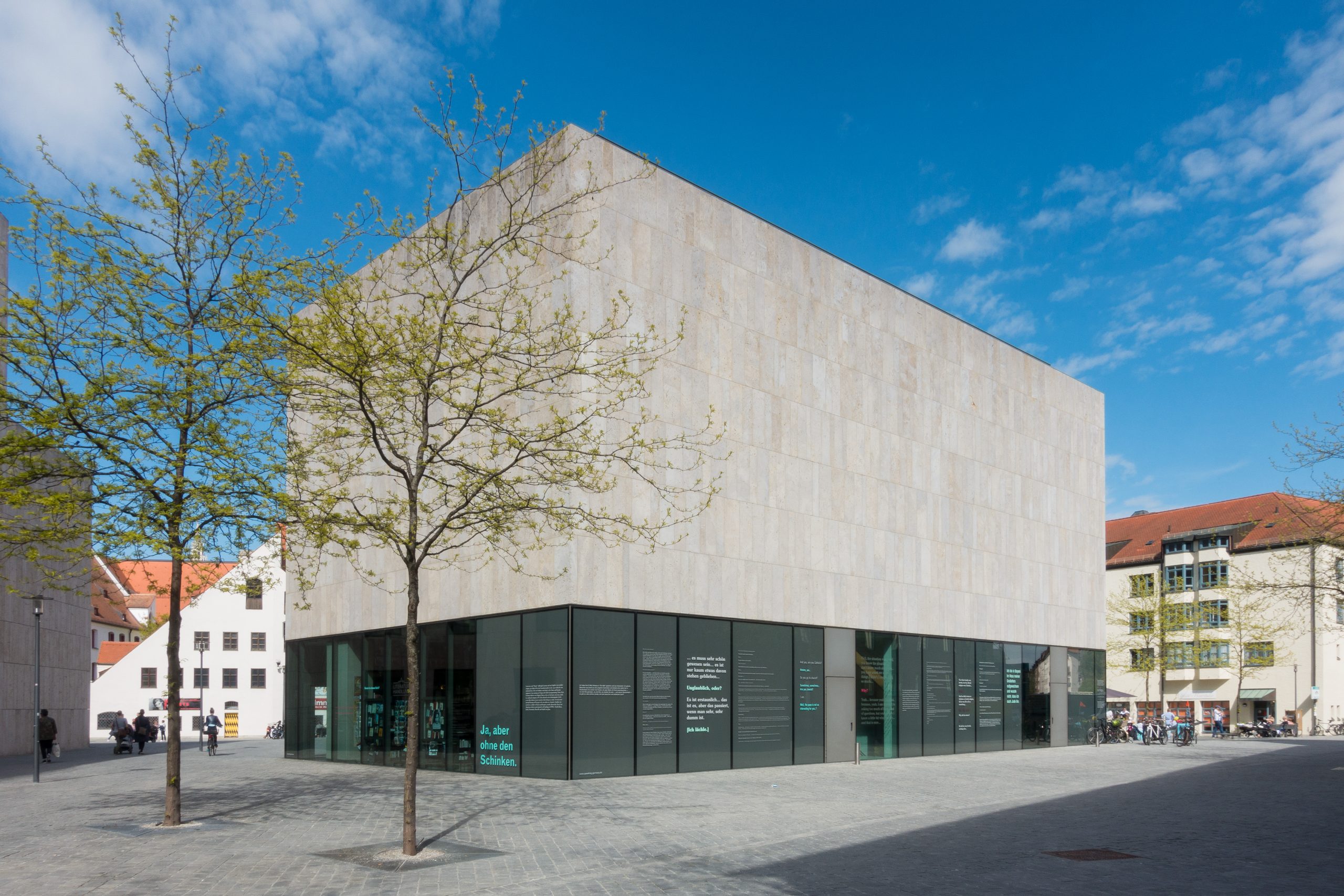 Das Jüdische Museum München ist in einer Außenansicht bei schönem Wetter zu sehen, vorne links zwei Bäume mit frischem Laub.